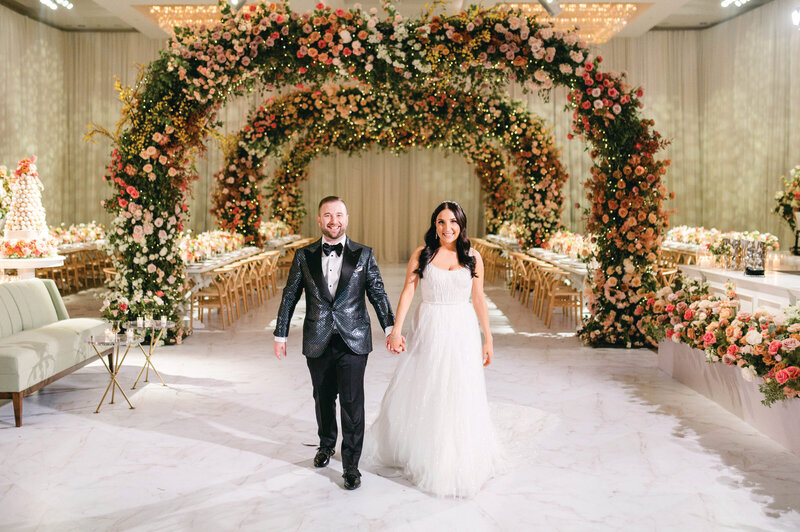Bride and groom walking hand in hand beneath an elaborate floral archway in an elegant ballroom