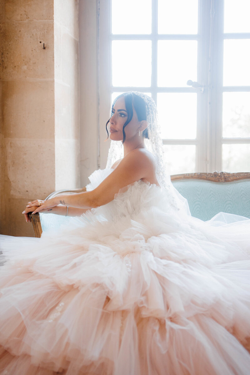 Bride seated in a voluminous pale blush wedding gown near a sunlit window