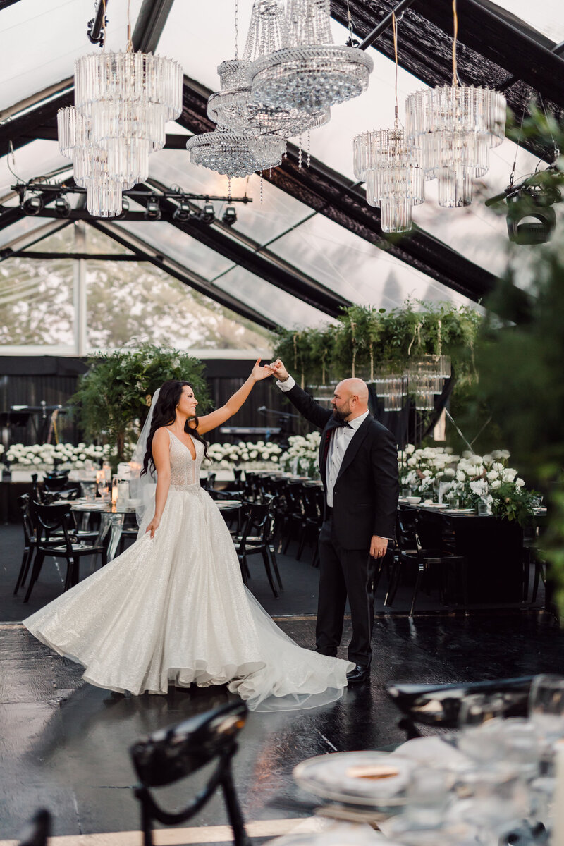 Bride and groom dancing beneath crystal chandeliers in a glass reception pavilion